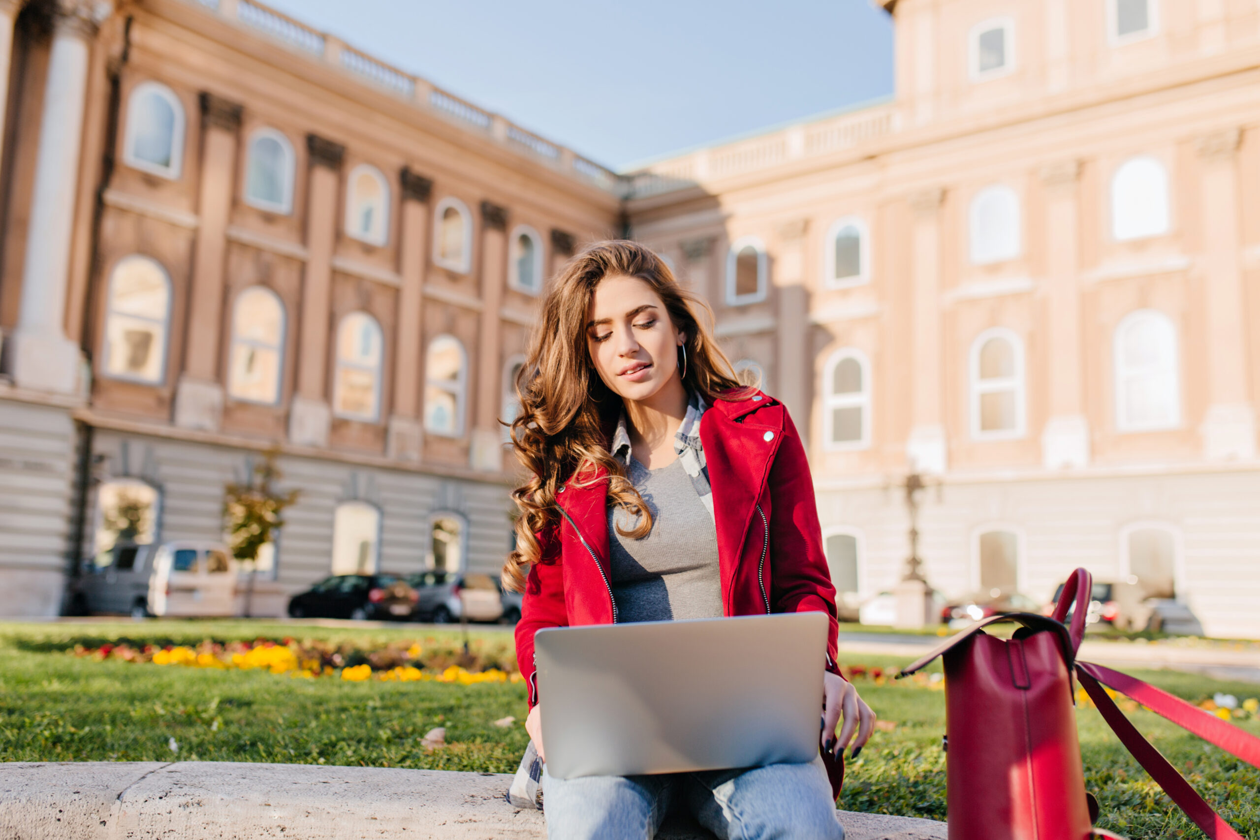 Outdoor portrait of serious curly female student sitting with laptop on the ground. Busy brunette girl in red jacket working with computer in front of college in warm day..