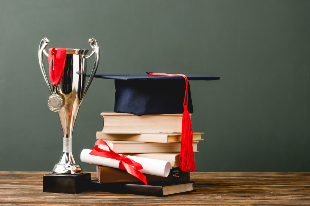 books, diploma, academic cap, trophy cup and medal on wooden surface isolated on grey