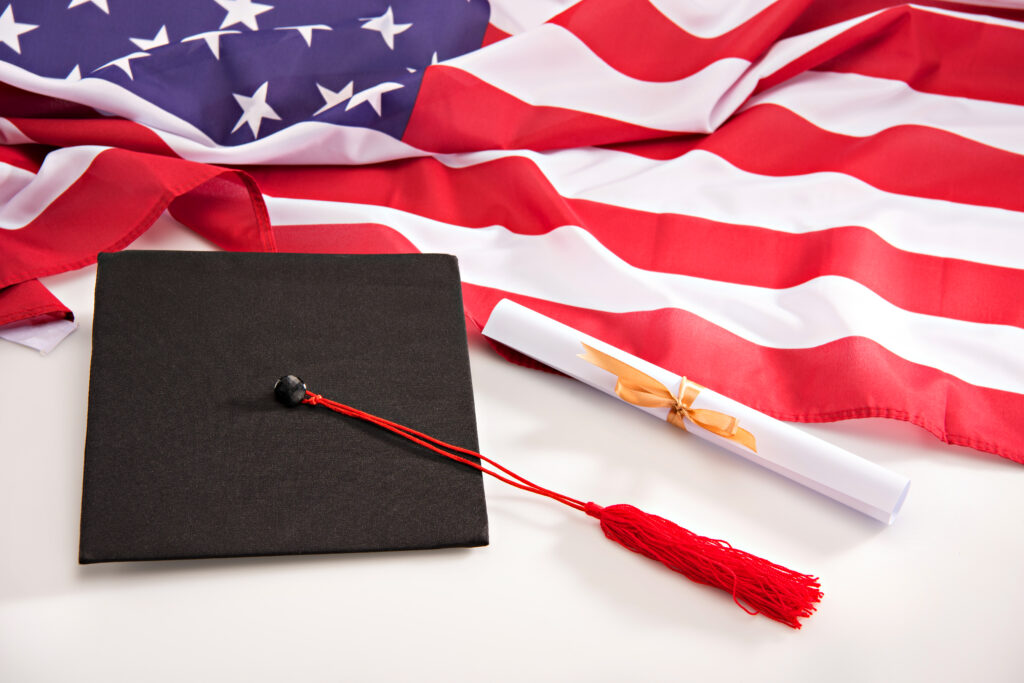 Close up view of graduation mortarboard, diploma and us flag on white, education concept
